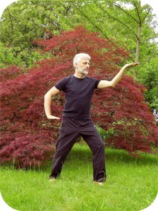 Un homme en tenue de tai chi effectue une pose équilibrée, les bras étendus et les jambes légèrement fléchies, dans une attitude de concentration et de sérénité.